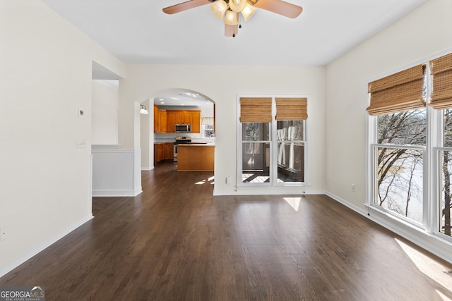 interior space featuring arched walkways, a wainscoted wall, dark wood finished floors, a ceiling fan, and baseboards