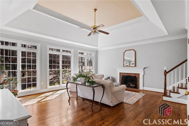 living area with a raised ceiling, ornamental molding, a brick fireplace, wood finished floors, and stairs