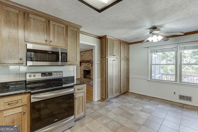kitchen featuring stainless steel appliances, a ceiling fan, crown molding, dark stone countertops, and a wainscoted wall