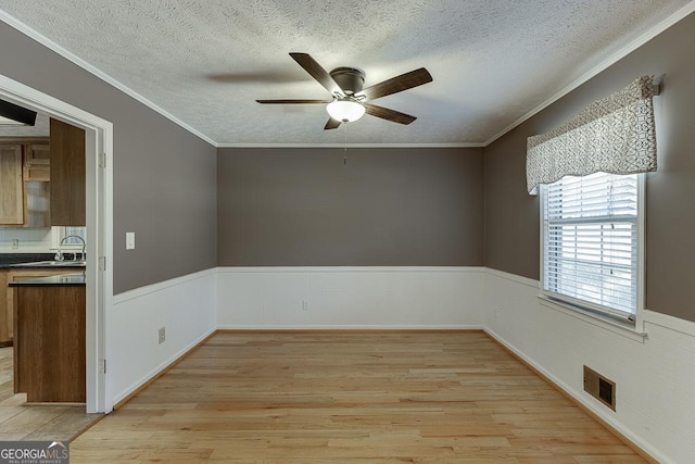 spare room with ceiling fan, light wood finished floors, crown molding, wainscoting, and a textured ceiling