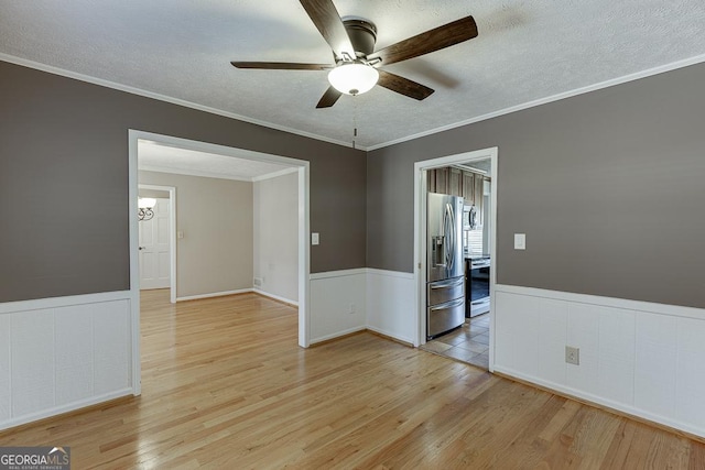 empty room featuring a ceiling fan, light wood-style flooring, a textured ceiling, a wainscoted wall, and ornamental molding