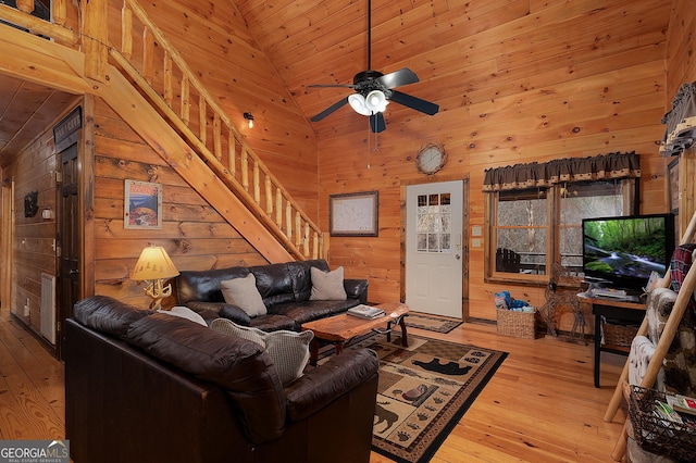 living area with light wood-style flooring, stairway, a ceiling fan, wooden walls, and high vaulted ceiling