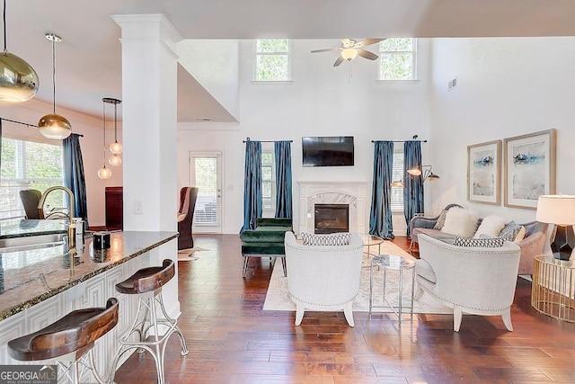 living room with decorative columns, dark wood finished floors, a ceiling fan, a towering ceiling, and a high end fireplace