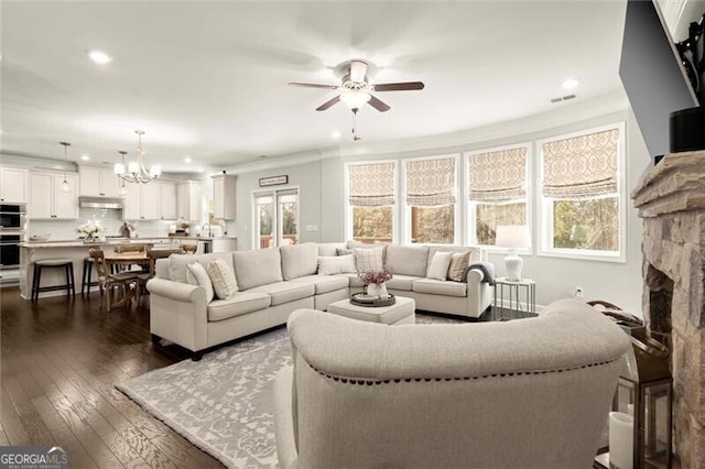 living room featuring dark wood-style floors, recessed lighting, ornamental molding, a stone fireplace, and ceiling fan with notable chandelier