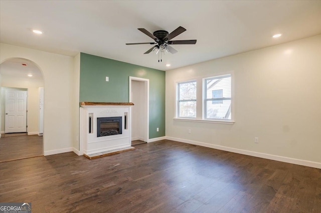 unfurnished living room with dark wood-style floors, arched walkways, a ceiling fan, a glass covered fireplace, and baseboards