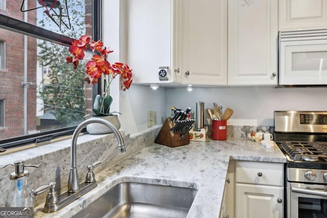 kitchen featuring stainless steel gas range oven, white cabinets, white microwave, light stone countertops, and a sink