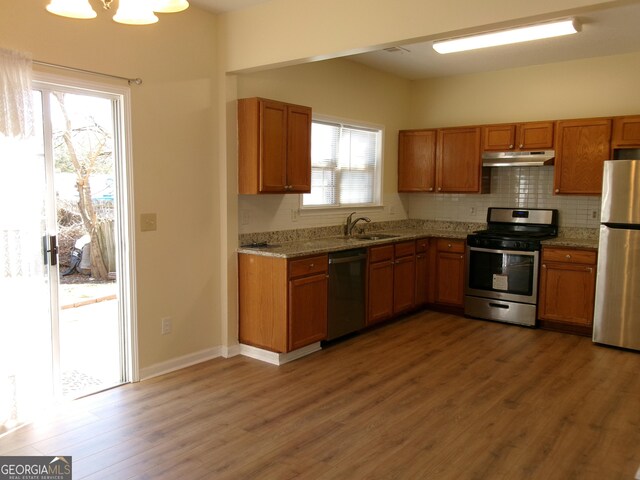 kitchen with brown cabinetry, wood finished floors, stainless steel appliances, under cabinet range hood, and backsplash