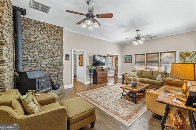 living area with dark wood-type flooring, visible vents, a ceiling fan, ornamental molding, and a wood stove