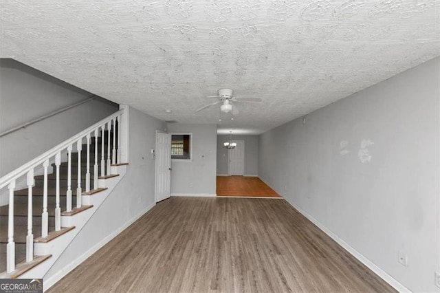 unfurnished living room with ceiling fan with notable chandelier, a textured ceiling, wood finished floors, baseboards, and stairs