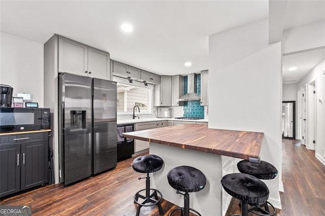 kitchen featuring black appliances, wall chimney exhaust hood, a kitchen bar, and wooden counters