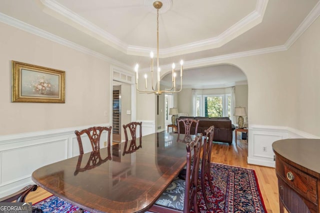 dining space featuring arched walkways, a wainscoted wall, a notable chandelier, a raised ceiling, and light wood-type flooring