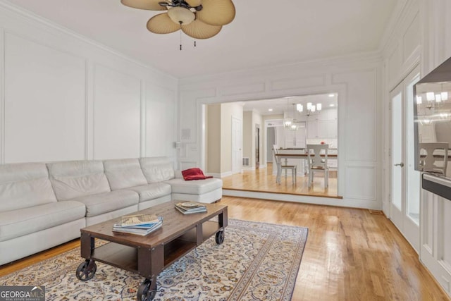 living area with ornamental molding, light wood-type flooring, a decorative wall, and ceiling fan with notable chandelier