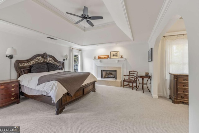 carpeted bedroom featuring a tray ceiling, arched walkways, crown molding, a fireplace with raised hearth, and a ceiling fan