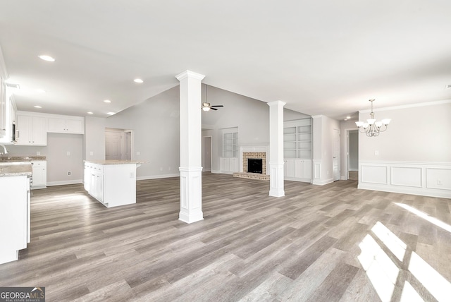 unfurnished living room featuring decorative columns, light wood-style floors, vaulted ceiling, a brick fireplace, and ceiling fan with notable chandelier