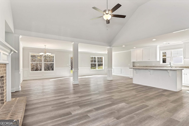 unfurnished living room featuring light wood-style flooring, vaulted ceiling, a sink, ornate columns, and ceiling fan with notable chandelier