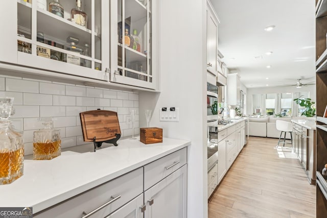 kitchen featuring ceiling fan, open floor plan, backsplash, light wood finished floors, and glass insert cabinets