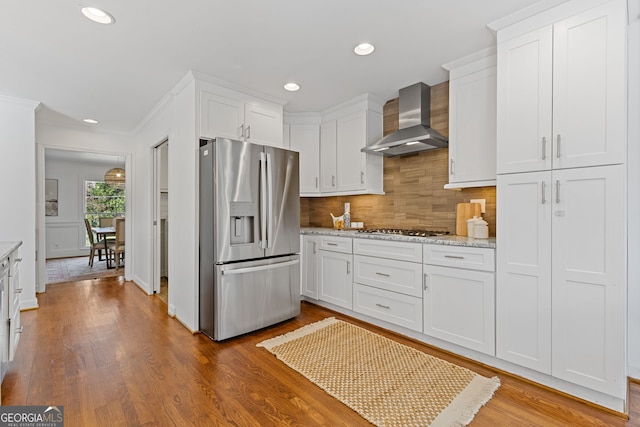 kitchen featuring wood finished floors, appliances with stainless steel finishes, white cabinetry, wall chimney range hood, and backsplash