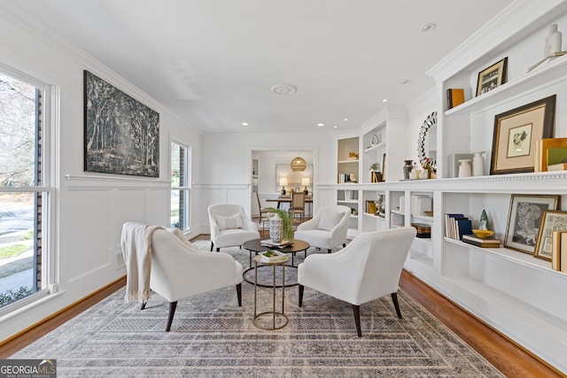 sitting room featuring built in shelves, wood finished floors, a wainscoted wall, ornamental molding, and a decorative wall