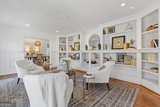 living area featuring built in shelves, wood finished floors, a wainscoted wall, crown molding, and a decorative wall