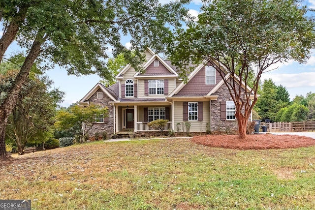 craftsman house featuring covered porch, stone siding, a front lawn, and fence