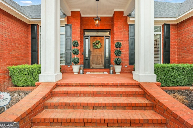 view of exterior entry featuring covered porch, brick siding, and roof with shingles