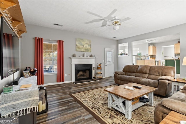 living room featuring a textured ceiling, a fireplace with raised hearth, wood finished floors, visible vents, and a ceiling fan