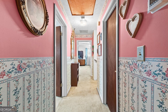 hallway featuring light carpet, a wainscoted wall, a textured ceiling, and attic access