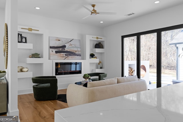 living area featuring visible vents, a glass covered fireplace, light wood-style floors, built in shelves, and recessed lighting