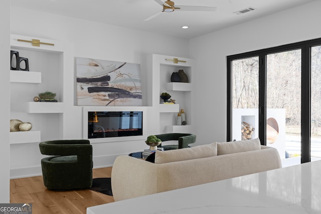 living area with ceiling fan, a glass covered fireplace, wood finished floors, and a healthy amount of sunlight