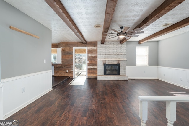 unfurnished living room featuring beam ceiling, a wainscoted wall, a fireplace, ceiling fan, and a textured ceiling