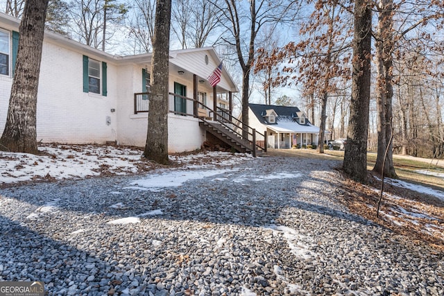 view of snowy exterior with gravel driveway, stairs, a porch, and brick siding