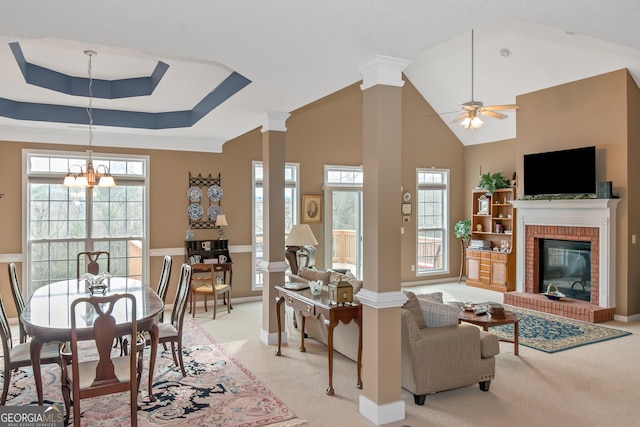 living room with light carpet, decorative columns, a raised ceiling, a brick fireplace, and ceiling fan with notable chandelier