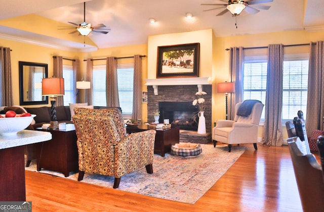 living area with ceiling fan, a stone fireplace, wood finished floors, and crown molding