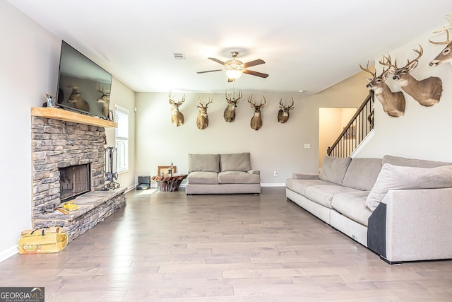 living area with stairs, ceiling fan, a stone fireplace, and wood finished floors