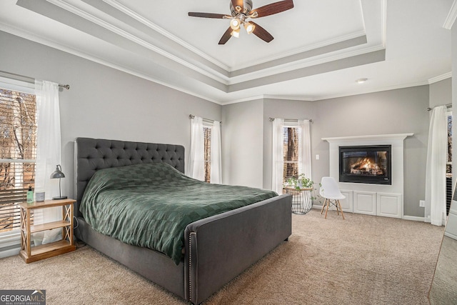 carpeted bedroom with a ceiling fan, a tray ceiling, crown molding, and a glass covered fireplace