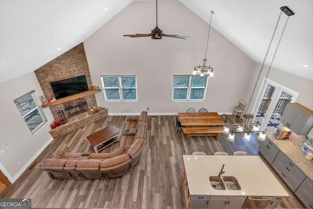 living room featuring high vaulted ceiling, a stone fireplace, ceiling fan with notable chandelier, wood finished floors, and baseboards