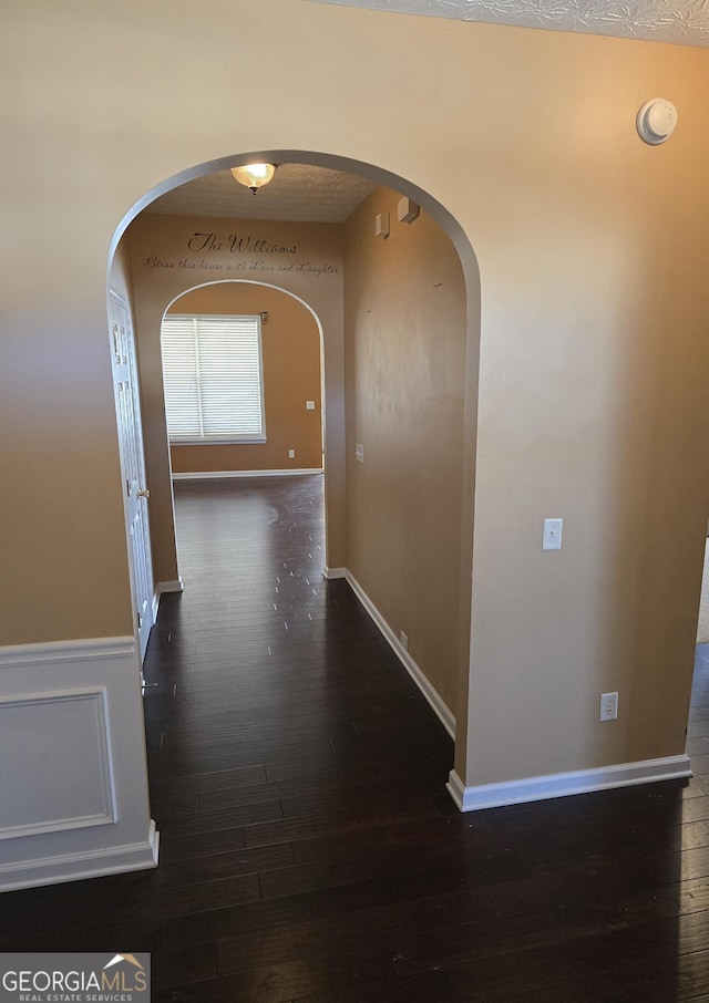 corridor with arched walkways, a wainscoted wall, wood-type flooring, and baseboards