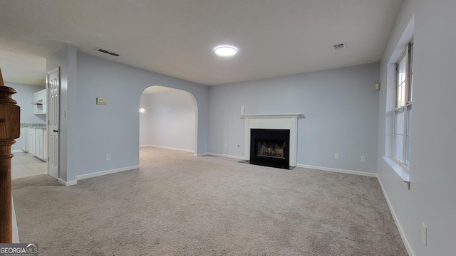 unfurnished living room with arched walkways, light colored carpet, visible vents, a fireplace with flush hearth, and baseboards