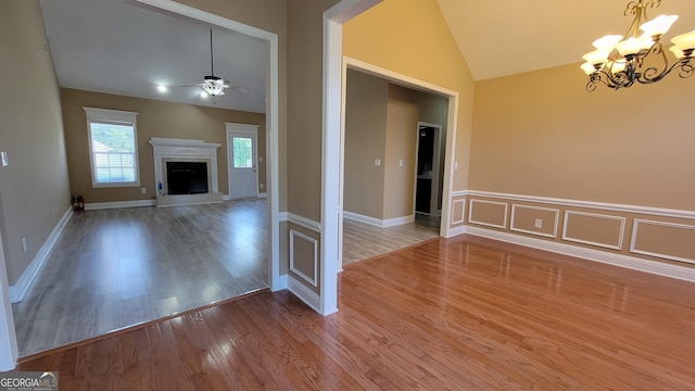 unfurnished living room with a fireplace with raised hearth, a decorative wall, vaulted ceiling, wood finished floors, and ceiling fan with notable chandelier