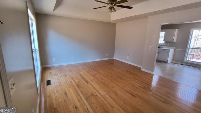 empty room with a tray ceiling, light wood finished floors, visible vents, ceiling fan, and baseboards