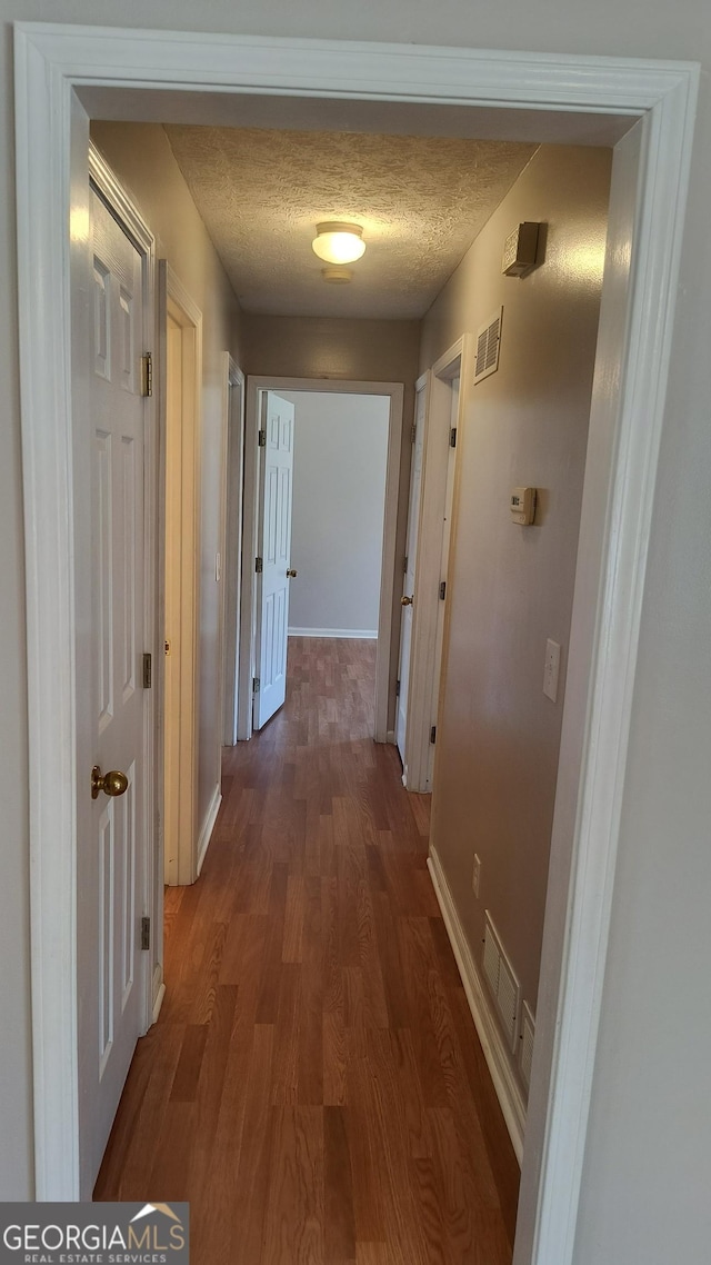 hallway with a textured ceiling, wood finished floors, visible vents, and baseboards