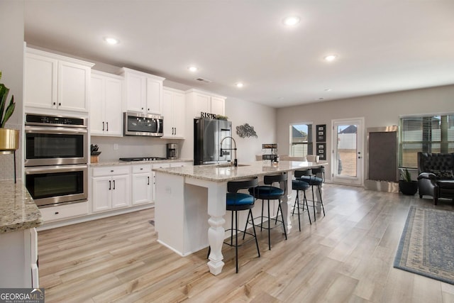 kitchen with white cabinets, an island with sink, light wood-style flooring, stainless steel appliances, and a sink