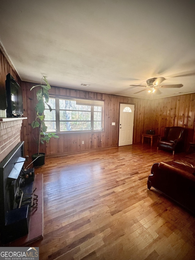 living room with ceiling fan, wood walls, wood finished floors, visible vents, and a brick fireplace