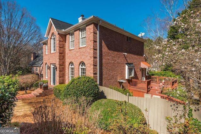 view of property exterior with a deck, brick siding, a chimney, and fence