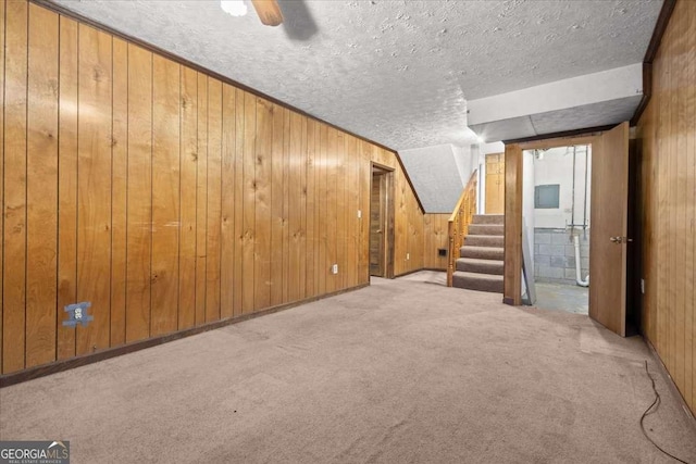 finished basement with wooden walls, a ceiling fan, stairs, a textured ceiling, and carpet floors