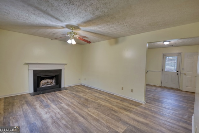 unfurnished living room with a textured ceiling, a fireplace with flush hearth, wood finished floors, a ceiling fan, and baseboards