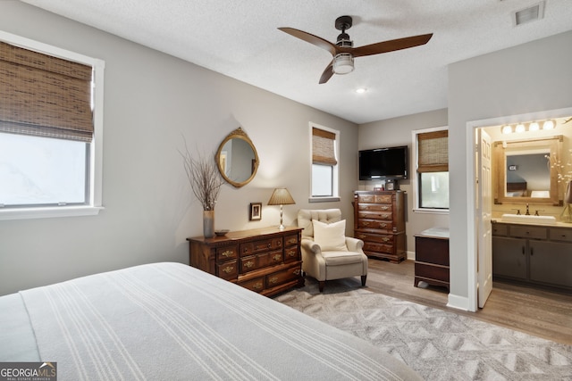 bedroom featuring light wood finished floors, visible vents, ensuite bathroom, a sink, and a textured ceiling