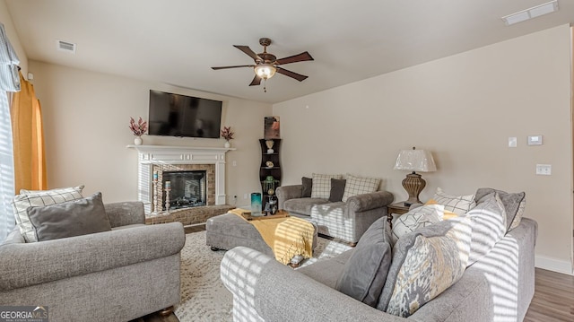 living room with a brick fireplace, a ceiling fan, visible vents, and wood finished floors