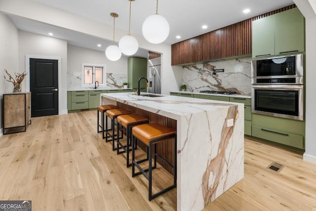 kitchen featuring light wood finished floors, a center island with sink, green cabinetry, stainless steel appliances, and a sink