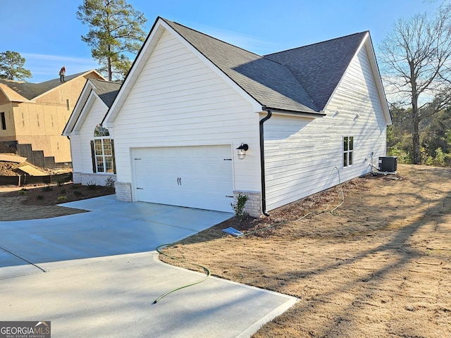 view of side of home with a garage, concrete driveway, roof with shingles, and central air condition unit